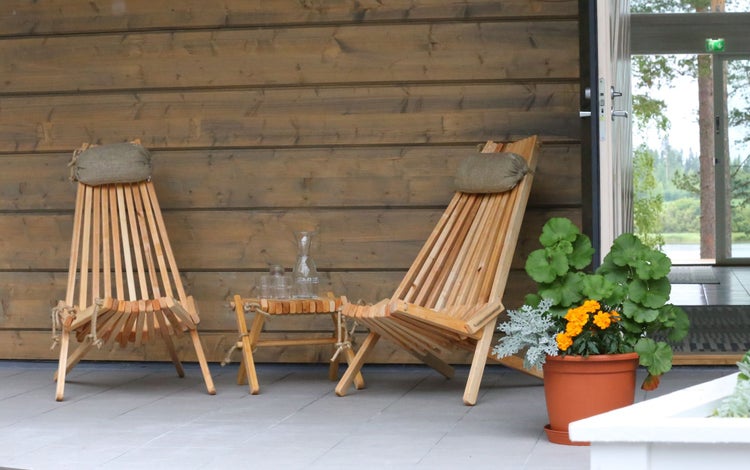 Zwei Gartenliegen aus Holzlamellen mit Kopfkissen und passendem Beistelltisch auf einer Terrasse vor einer Holzwand mit einer Topfpflanze.