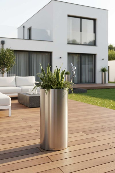 Hoher zylindrischer Pflanzkübel aus Edelstahl auf einer modernen Holzterrasse mit Gartenmöbeln und einem Wohnhaus im Hintergrund.