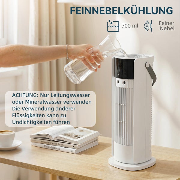 Weißer Turmventilator mit Feinnebelkühlung wird mit Wasser aus einer Karaffe befüllt. Das Gerät steht auf einem hellen Holztisch neben Büchern.