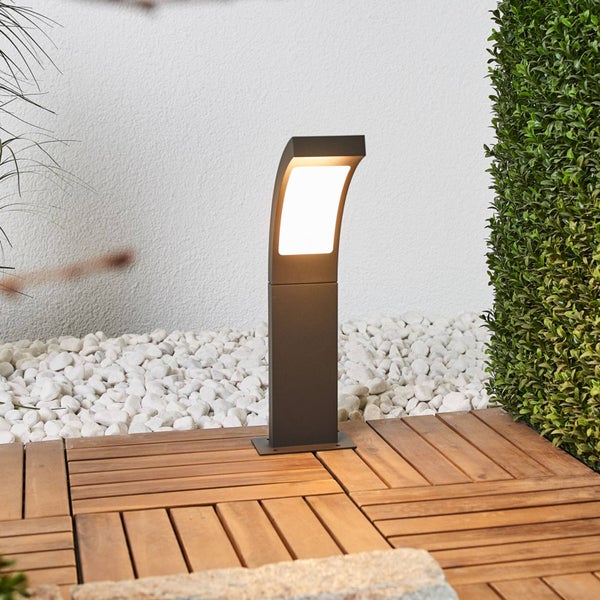 Moderne LED-Pollerleuchte auf einer Holzterrasse neben weißen Ziersteinen und einer grünen Hecke.