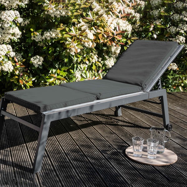 Moderne Gartenliege mit dunkler Auflage und Rollen auf einer Holzterrasse vor einer blühenden Hecke.