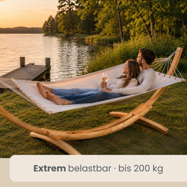 Zwei Personen entspannen in einer hellen Hängematte mit stabilem Holzgestell an einem Seeufer. Belastbarkeit bis 200 Kilogramm.