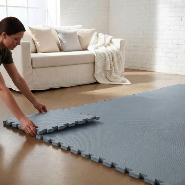 Eine Frau verlegt graue Puzzlematten aus Schaumstoff in einem hellen, organisierten Wohnzimmer mit weißem Sofa und Ziegelwand.