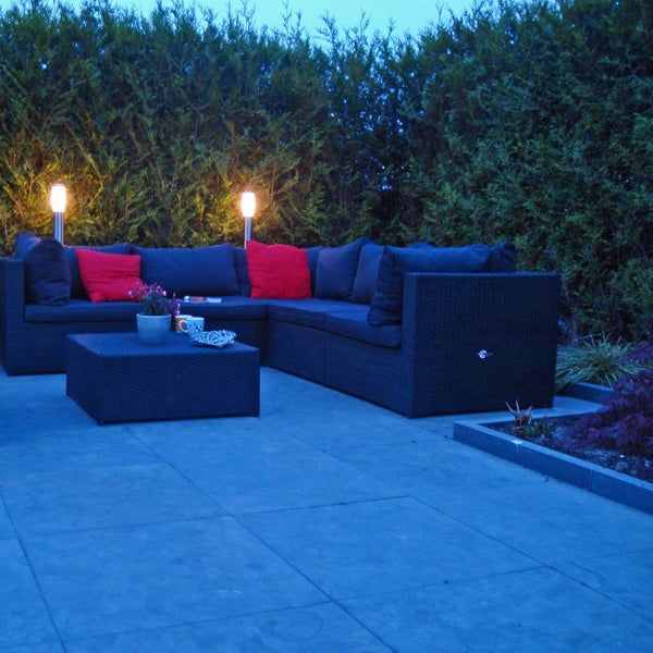 Outdoor-Lounge-Set aus dunklem Polyrattan in L-Form auf einer Terrasse mit passendem Couchtisch, roten Kissen und Gartenbeleuchtung.