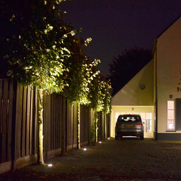Einfahrt bei Nacht mit Bodeneinbaustrahlern, die Bäume entlang eines Holzzauns neben einem Haus und einem Auto beleuchten.