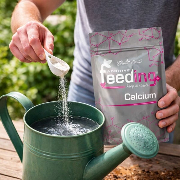 Eine Person fügt Calciumdünger aus einem Messlöffel in eine Gießkanne. Im Hintergrund ist die Verpackung des Green House Additive Feeding Calcium Düngers zu sehen.