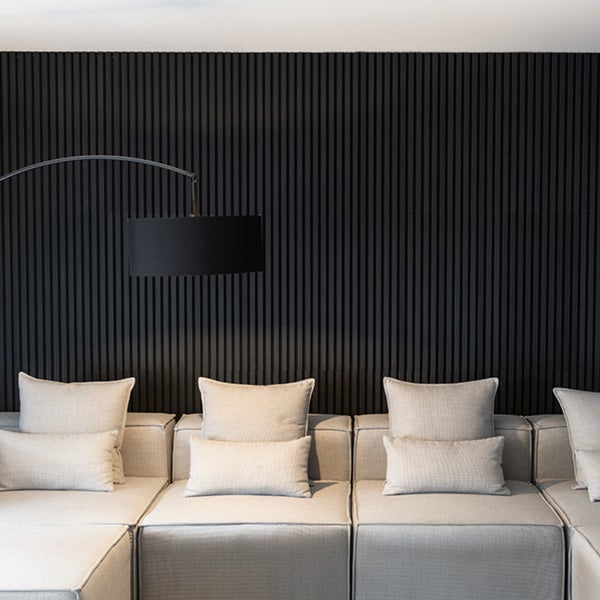 Akustikpaneele aus schwarzen Holzlamellen an einer Wohnzimmerwand hinter einem hellgrauen Sofa mit einer schwarzen Stehlampe.