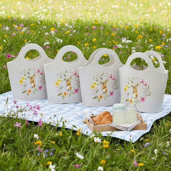 Vier Filztaschen mit Osterhasen und Blumenmotiv auf einer Picknickdecke im Blumenwiese mit Croissants und Getränken in Einmachgläsern.