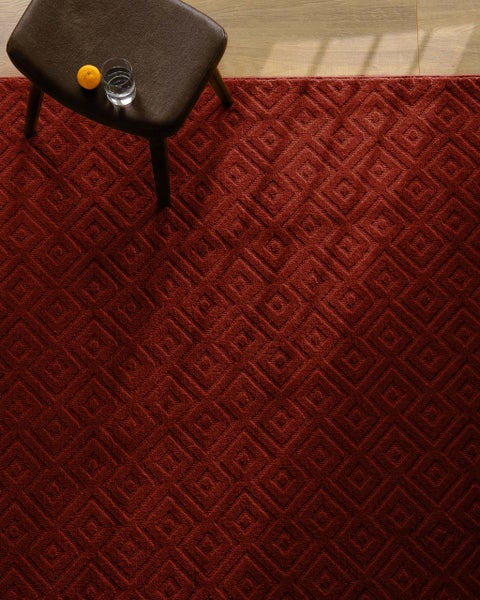 Strukturierter Teppich mit geometrischem Rautenmuster auf hellem Holzboden, neben einem gepolsterten Hocker.