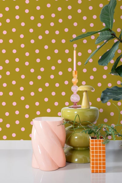 Vliestapete mit rosa Punkten auf olivfarbenem Untergrund in einer modernen Raumszene mit Designmöbeln und Dekovase.