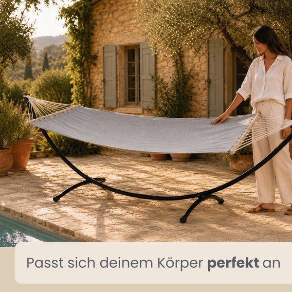 Hängematte mit schwarzem Metallgestell auf einer Steinterrasse am Pool vor einem Steinhaus. Eine Frau steht daneben.