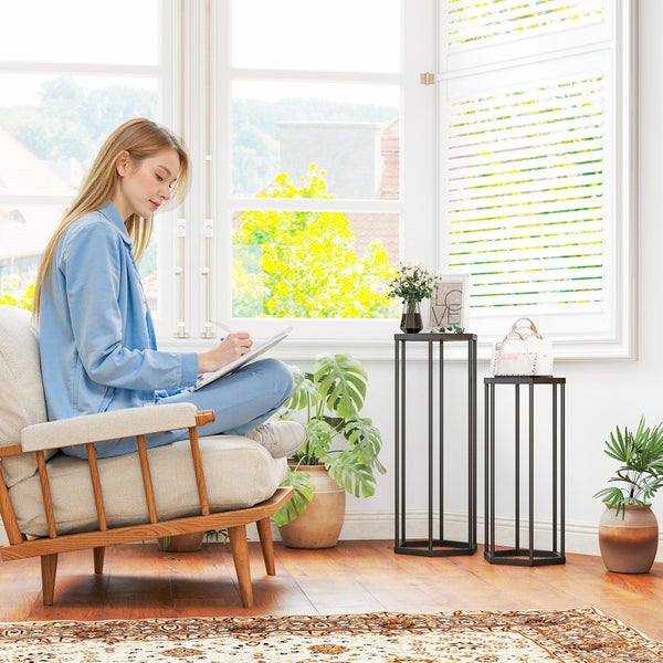 Modernes Wohnzimmer mit zwei schwarzen Pflanzensäulen aus Metall in unterschiedlichen Höhen, hellem Sessel und Zimmerpflanzen vor einem Fenster.