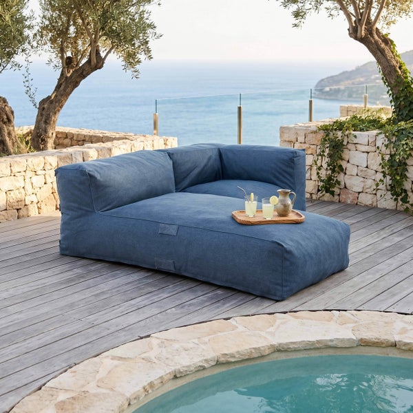 Blaues Outdoor-Lounge-Eckmodul auf einer Holzterrasse mit Steinmauer und Meerblick.