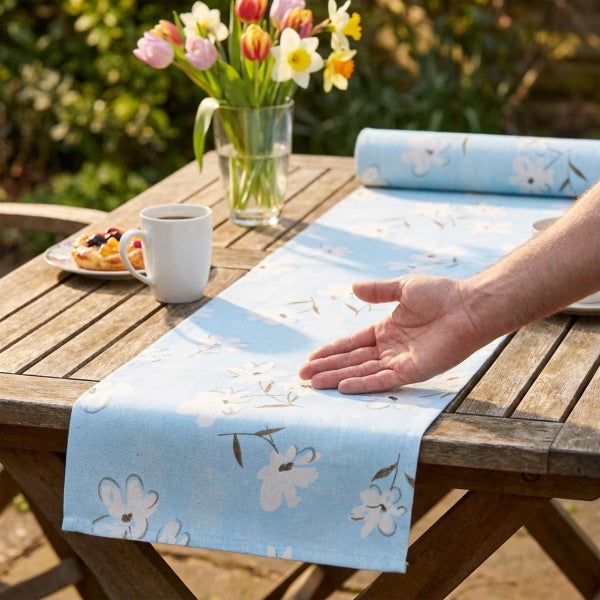 Tischläufer mit Blumenmuster auf einem Holztisch im Freien, präsentiert von einer Hand.