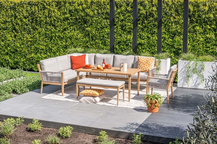 Modernes Garten-Lounge-Set aus Holz mit Ecksofa, Tisch, Bank und Sessel auf einer grauen Terrasse vor einer grünen Hecke.