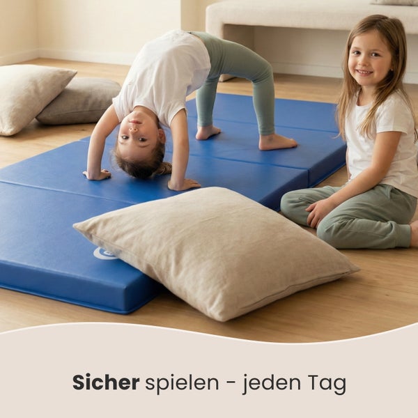 Zwei Kinder spielen auf einer blauen Turnmatte mit Kissen in einem hellen Raum.