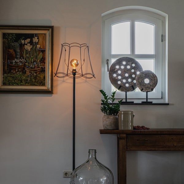 Stehleuchte mit Drahtschirm und Filament-Leuchtmittel in rustikalem Wohnraum neben Holztisch und Fenster mit Dekoration.