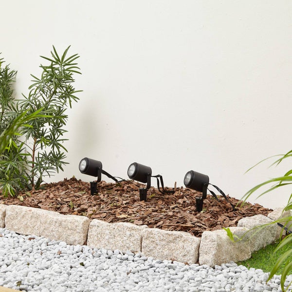 Drei schwarze Gartenstrahler mit Erdspießen in einem mit Mulch gefüllten Gartenbeet, eingefasst von hellen Natursteinen.