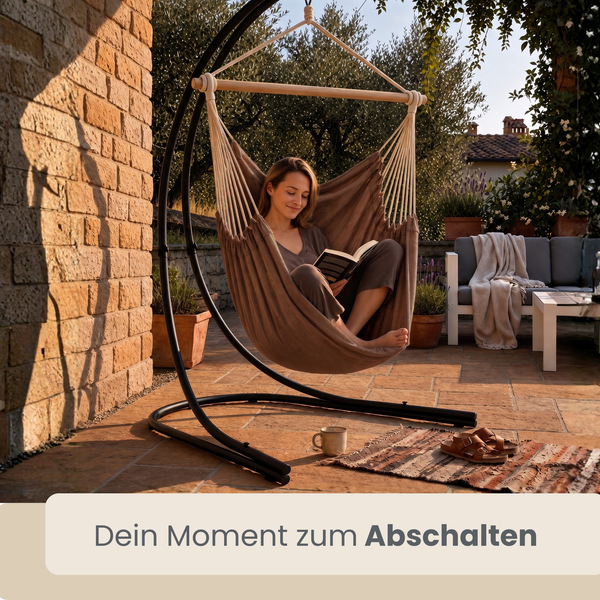 Frau sitzt lesend in einem Hängesessel mit Gestell auf einer Terrasse im Garten.