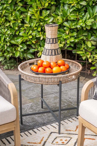 Runder Beistelltisch für den Außenbereich mit geflochtener Tischplatte und Metallgestell, dekoriert mit Vase und Obstschale auf einer Terrasse.
