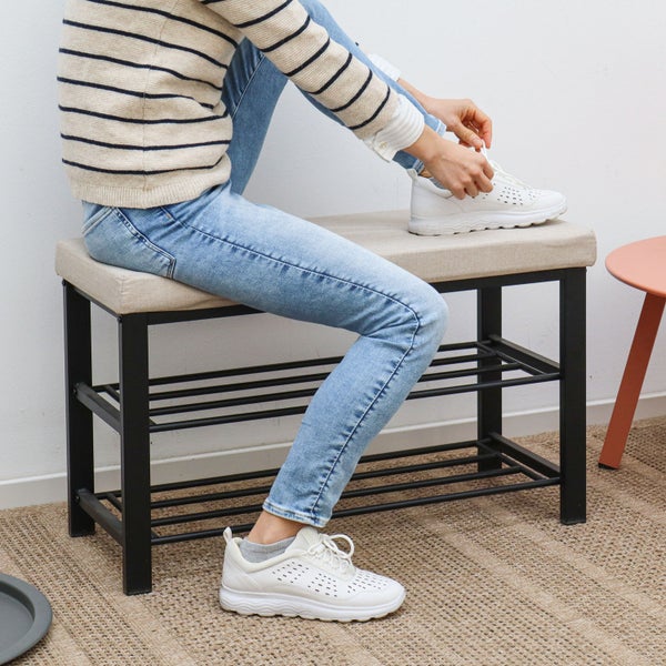 Schuhbank mit schwarzem Metallgestell, zwei Ablagen für Schuhe und hellbeiger gepolsterter Sitzfläche, auf der eine Person sitzt.