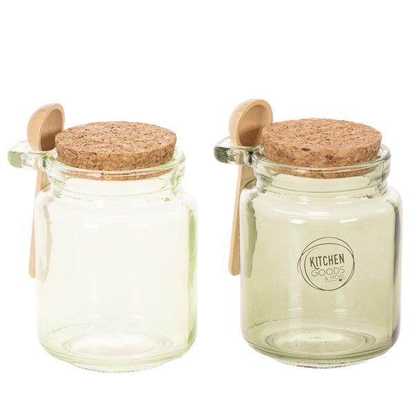 Zwei Vorratsgläser aus Glas mit Korkdeckeln und Holzlöffeln. Ein Glas mit Kitchen Goods and More Logo.