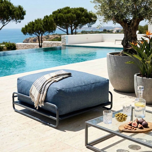 Moderner blauer Outdoor-Pouf mit grauem Metallgestell auf einer hellen Terrasse neben einem Pool mit Meerblick und großen Pflanzkübeln.
