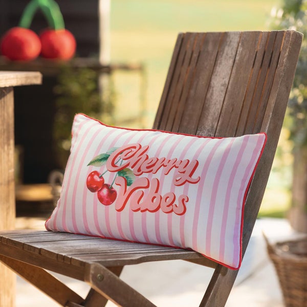Kissen mit Cherry Vibes Schriftzug, Kirschmotiv und Streifen auf einem Gartenstuhl aus Holz.