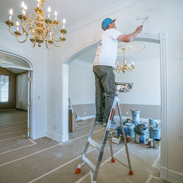 Ein Maler auf einer Trittleiter streicht die Wand über einem Rundbogen in einem hellen, großzügigen Innenraum während einer Renovierung.