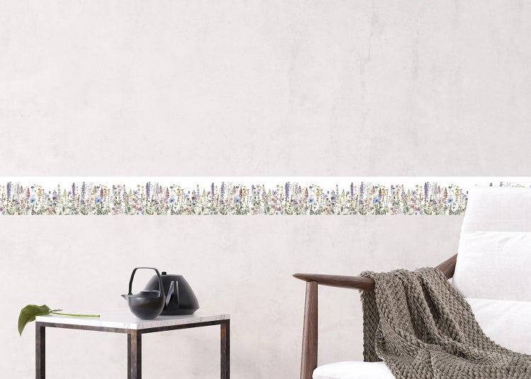 Selbstklebende Wandbordüre mit Wiesenblumen-Motiv in einem hellen Wohnbereich hinter einem Sofa mit strukturiertem Kissen und einer Obstschale.
