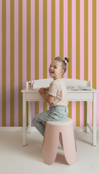 Gestreifte Tapete in Gold und Rosa in einem hellen Kinderzimmer mit weißem Schreibtisch und rosa Hocker.