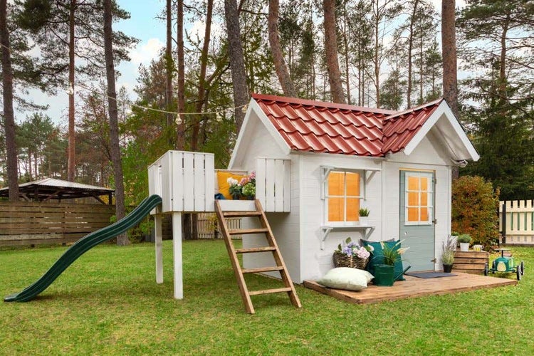 Weißes Holzspielhaus mit rotem Satteldach, grüner Tür, Fenster, erhöhter Plattform, grüner Rutsche und Holzleiter auf einer Rasenfläche im Garten.