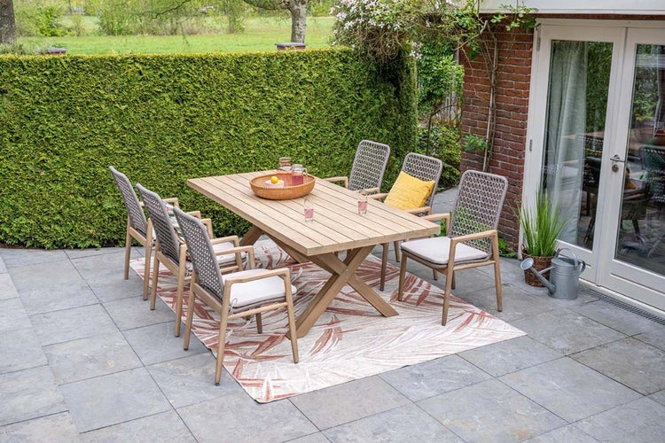 Moderne Garten-Essgruppe mit großem Holztisch und sechs Stühlen mit Geflecht-Rückenlehnen auf einem gemusterten Outdoor-Teppich auf einer Terrasse.