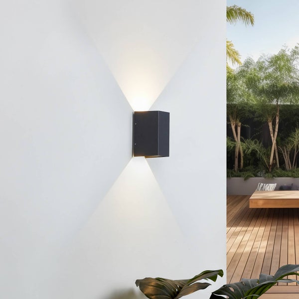 Rechteckige Außenwandleuchte in Schwarz an einer weißen Wand, die Licht nach oben und unten auf eine Holzterrasse mit Pflanzen wirft.