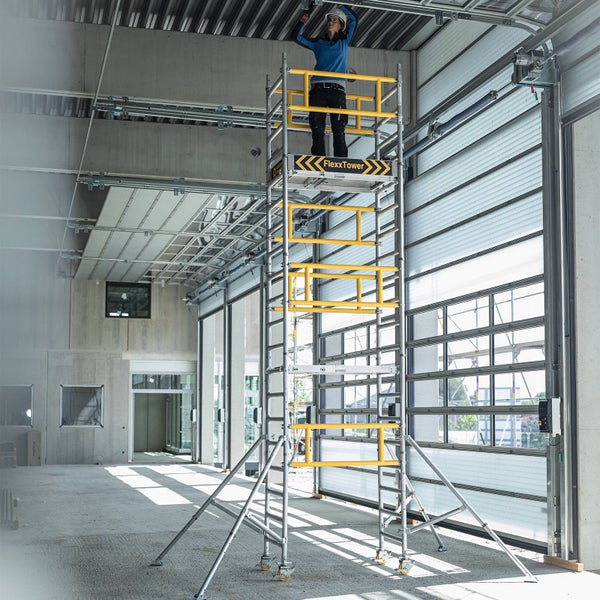 Person arbeitet an der Decke auf einem FlexxTower Aluminium-Fahrgerüst mit gelben Geländern und Auslegern in einer hellen Industriehalle.