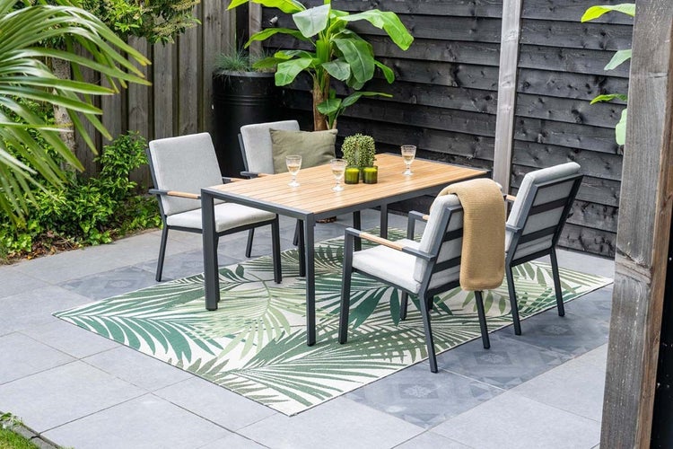 Gartenmöbel-Set mit einem Holztisch und vier gepolsterten Stühlen auf einem Außenteppich mit Blattmuster auf einer Terrasse.