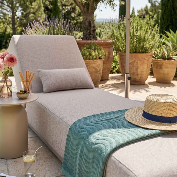 Gepolsterte Gartenliege mit Kissen, Handtuch und Strohhut auf einer hellen Terrasse mit mediterranen Pflanzen in Tontöpfen.