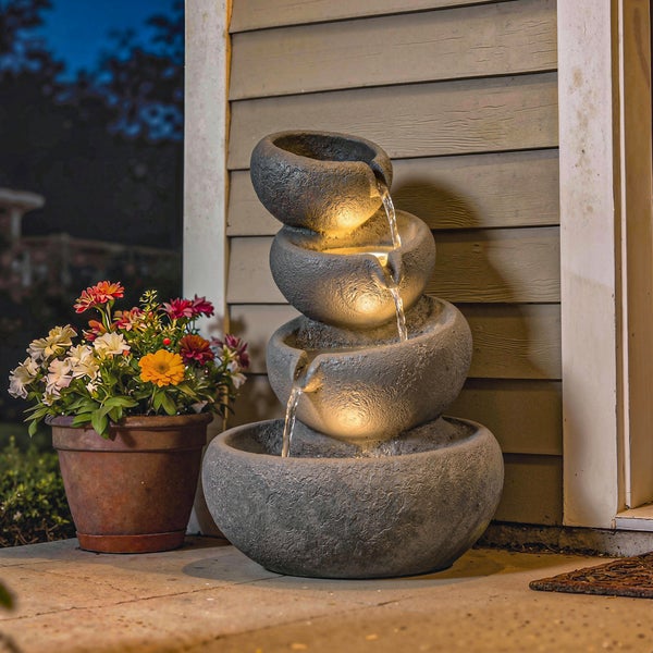 Gartenbrunnen in Steinoptik mit vier Etagen, warmweißer LED-Beleuchtung und fließendem Wasser in einer abendlichen Gartenlandschaft.