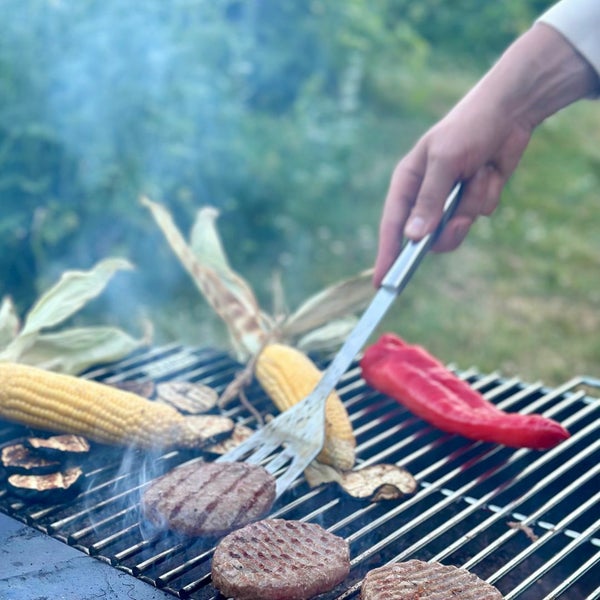 Eine Person grillt Fleischpattys, Maiskolben, Paprika und Auberginen mit einem Pfannenwender auf einem Grillrost aus Metall.