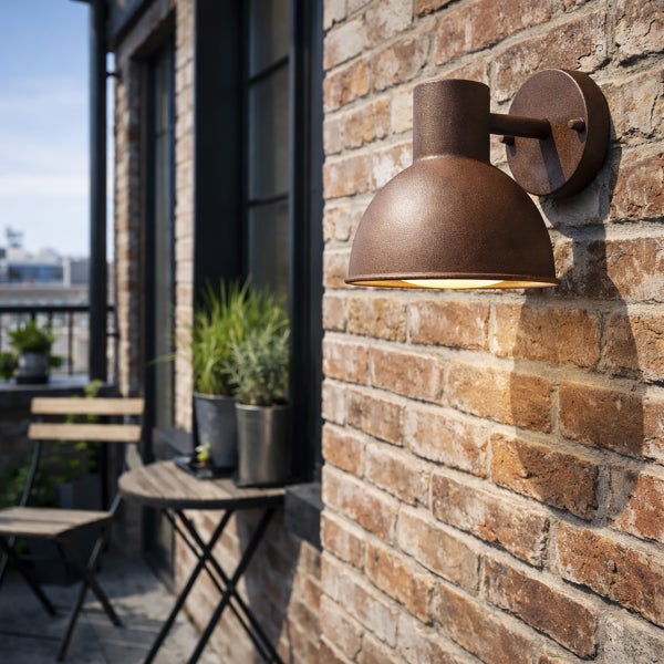 Außenwandleuchte in Rostoptik an einer Ziegelwand auf einem Balkon mit Tisch, Stuhl und Pflanzen.