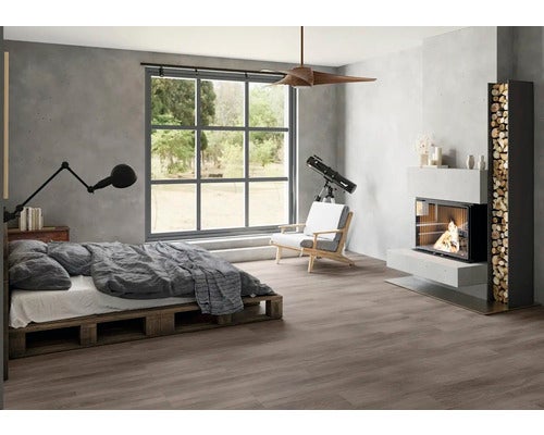 Geräumiges Zimmer mit dunklem Boden in Holzoptik, Palettenbett, Kamin mit Holzregal und großem Fenster im industriellen Stil.