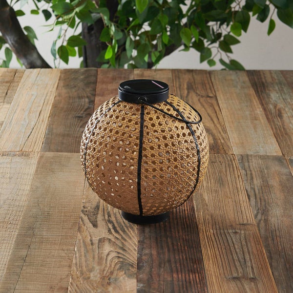 Runde Solar-Laterne in geflochtener Rattan-Optik mit schwarzem Metallrahmen auf einem rustikalen Holztisch.