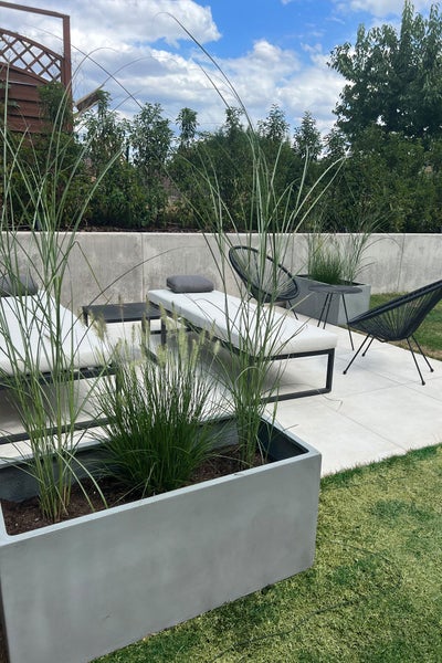 Moderne Terrasse mit großen grauen Pflanzkübeln in Betonoptik, hohen Ziergräsern, weißen Gartenliegen und schwarzen Loungesesseln.