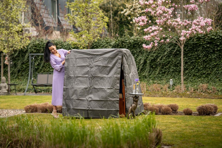 Gartenszene mit Frau, grauer Schutzhülle für Gartenmöbel und blühenden Bäumen