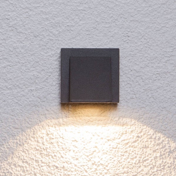 Quadratische Außenwandleuchte in Anthrazit mit Lichtaustritt nach unten auf einer strukturierten Fassade.