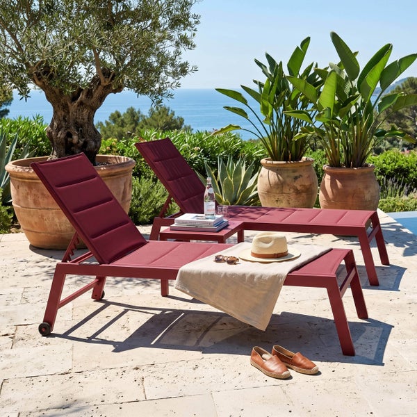 Zwei bordeauxrote Sonnenliegen aus Metall und Textilene auf einer Steinterrasse mit Pflanzen und Meerblick in sonniger Gartenatmosphäre.