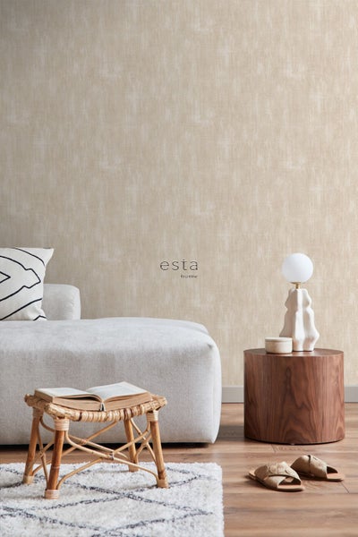 Vliestapete mit Textilstruktur von Esta home in einem hellen Wohnzimmer mit Sofa, Rattanhocker und Beistelltisch aus Holz. Esta home Logo.