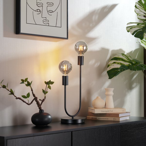 Moderne schwarze Tischleuchte aus Metall mit zwei Armen und Glühfaden-Leuchtmitteln auf einem Sideboard in einer hellen Wohnumgebung.