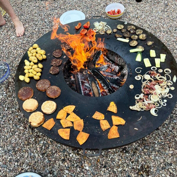 Kreisförmige schwarze Grillplatte über einer Feuerschale mit brennendem Holz, auf der Kartoffeln, Burger und Gemüse garen.