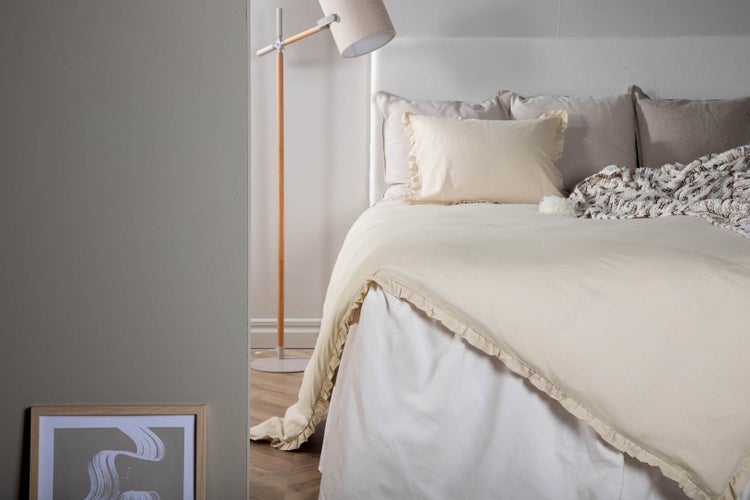 Helles Schlafzimmer mit Bettwäsche, Rüschen-Bettbezug, Stehlampe aus Holz und Bildrahmen auf dem Boden.